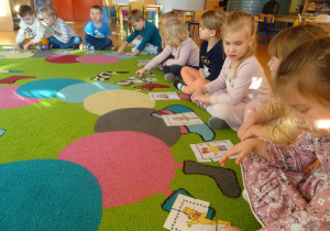Grupa dzieci siedzi na dywanie, każde z dzieci układa przed sobą obrazek z części przedstawiający misie.